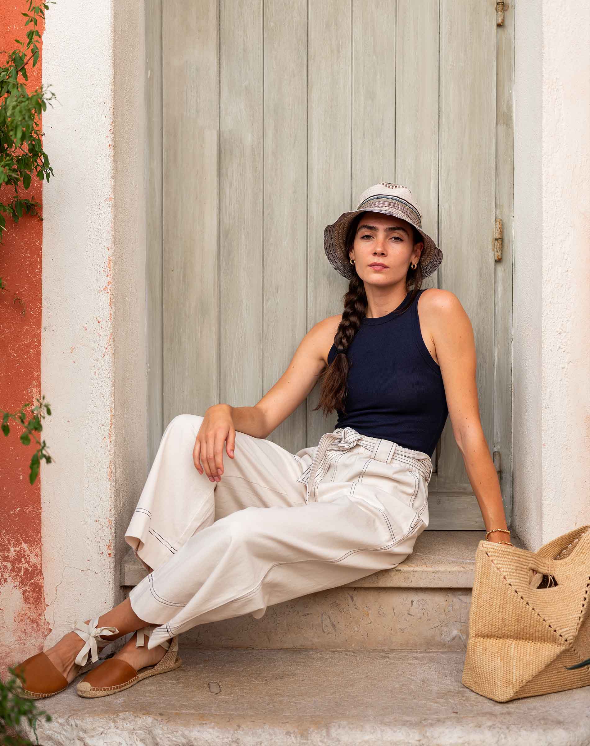Woman sitting on a step wearing a hat, navy top, and white pants with a straw bag.
