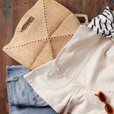Stack of folded clothes including a beige jacket, blue jeans, and a woven bag on a wooden surface.