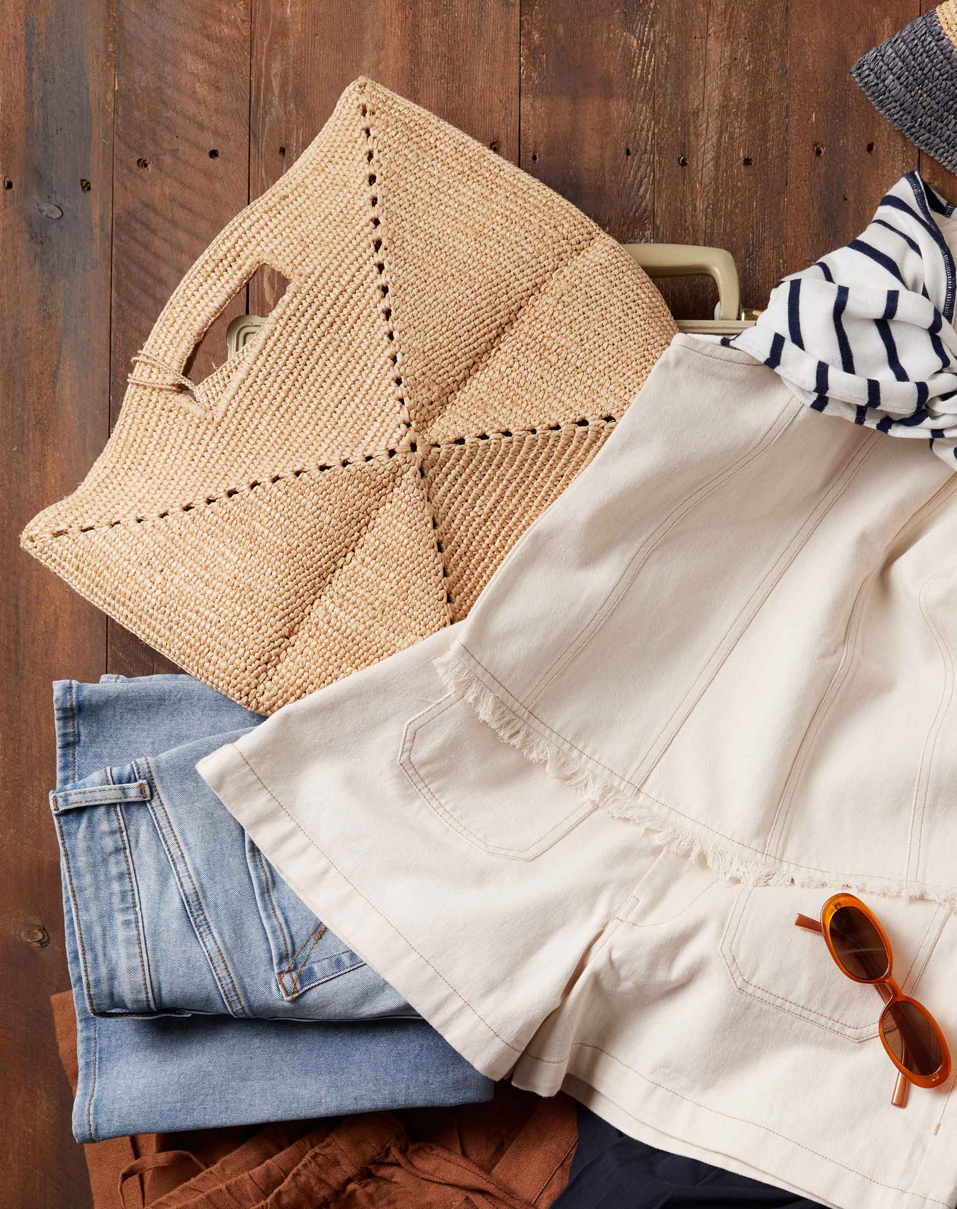 Stack of folded clothes including a beige jacket, blue jeans, and a woven bag on a wooden surface.