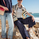 Two women sitting on rocks by the ocean, one wearing a striped cardigan and sunglasses.