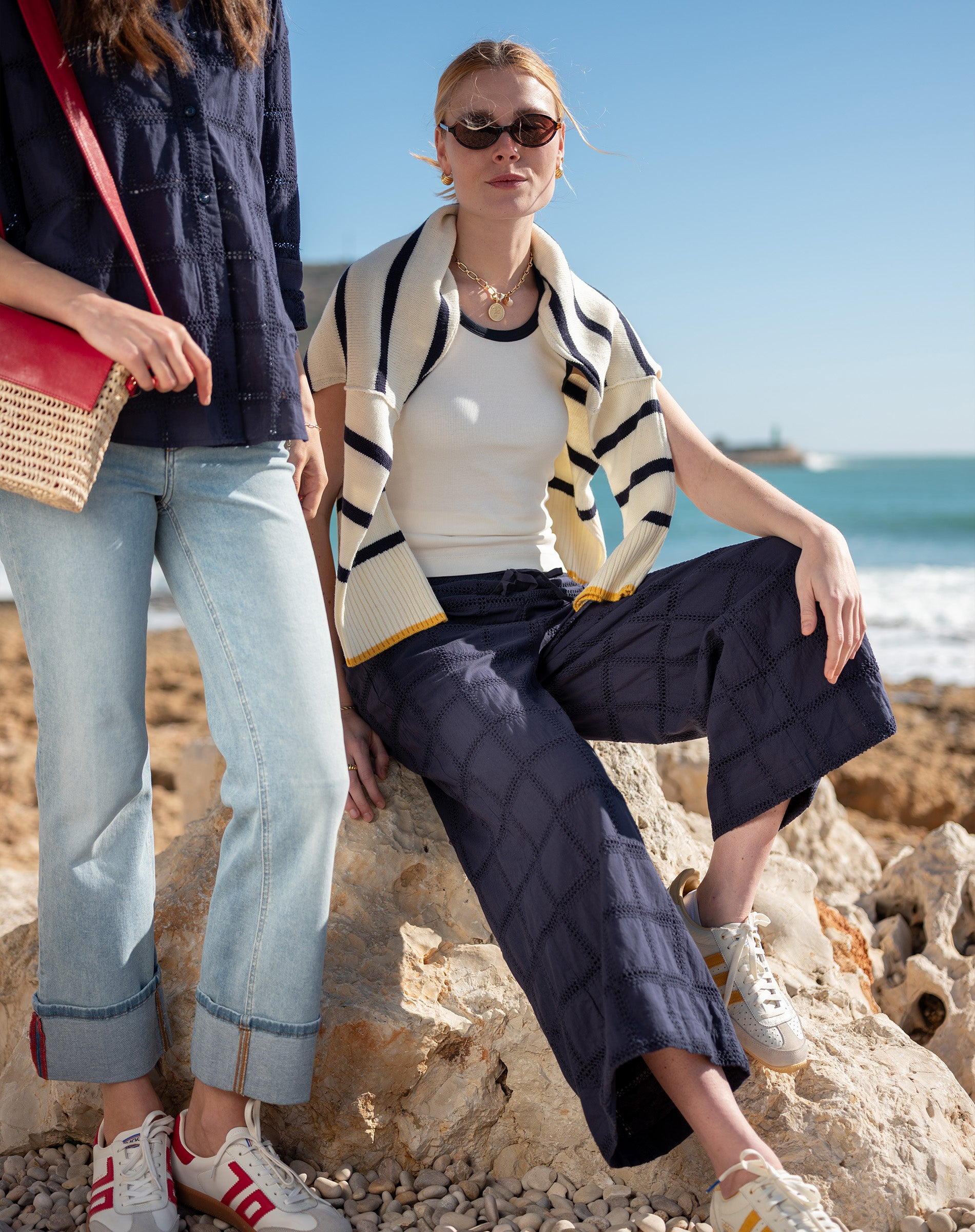 Two women sitting on rocks by the ocean, one wearing a striped cardigan and sunglasses.