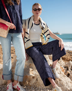 Two women sitting on rocks by the ocean, one wearing a striped cardigan and sunglasses.
