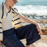 Woman sitting on rocks by the ocean wearing a striped sweater and navy pants.