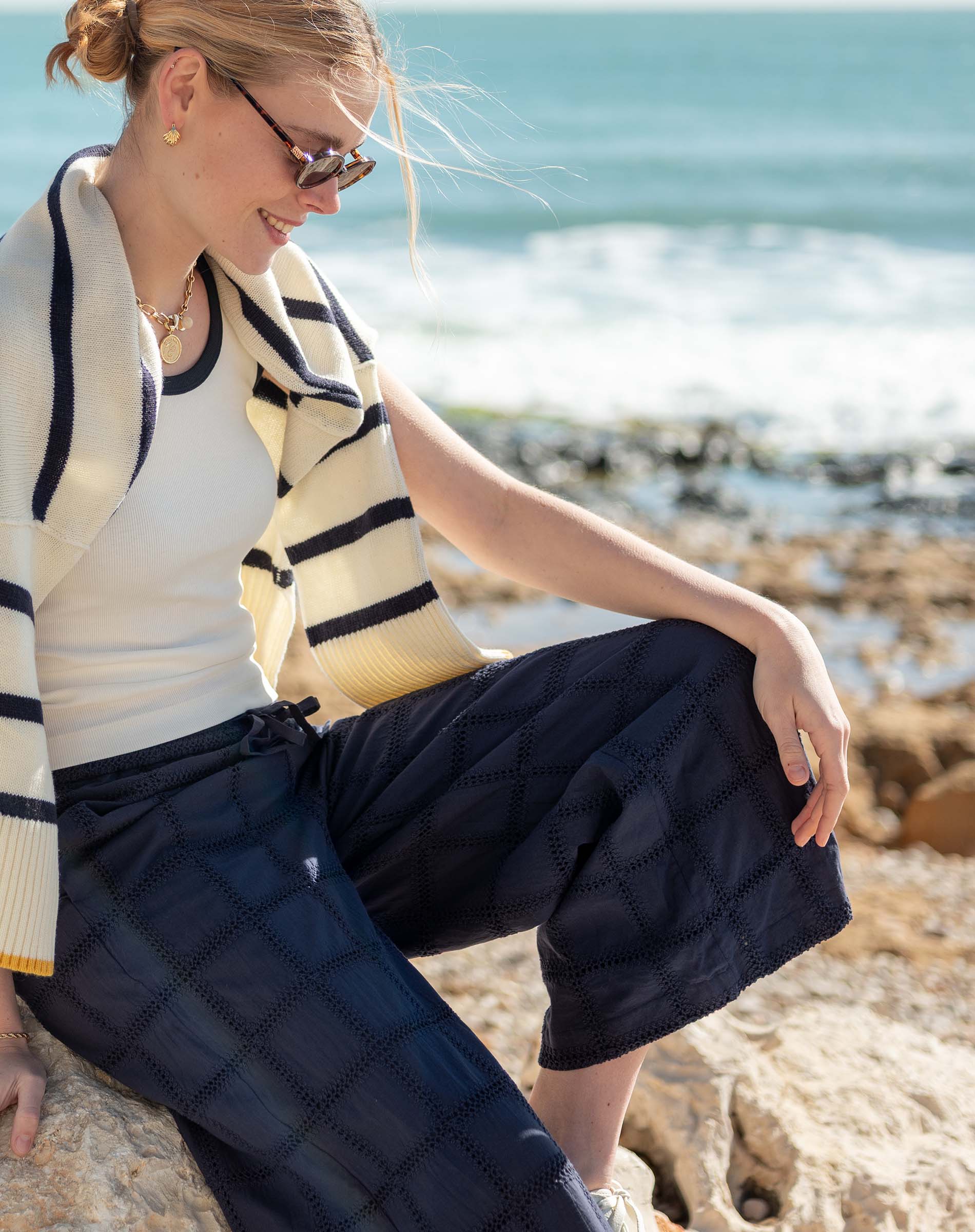 Woman sitting on rocks by the ocean wearing a striped sweater and navy pants.