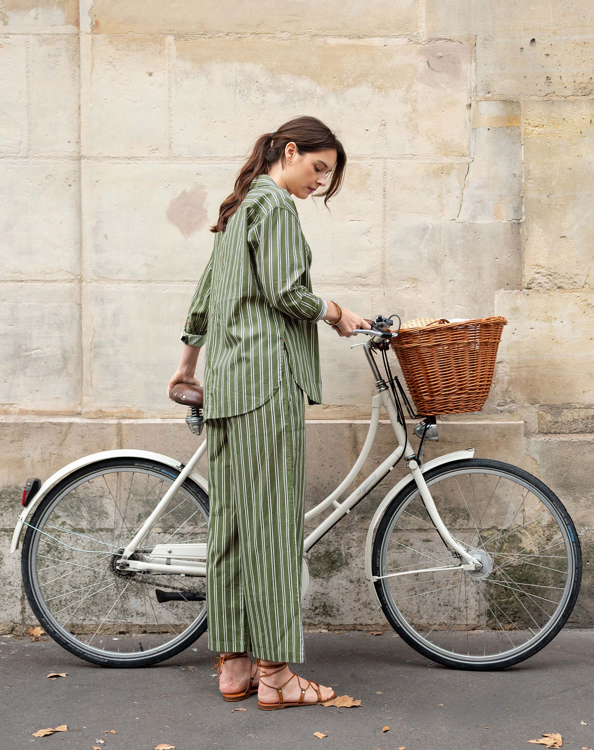 Woman in a green striped outfit standing next to a bicycle with a basket against a stone wall.
