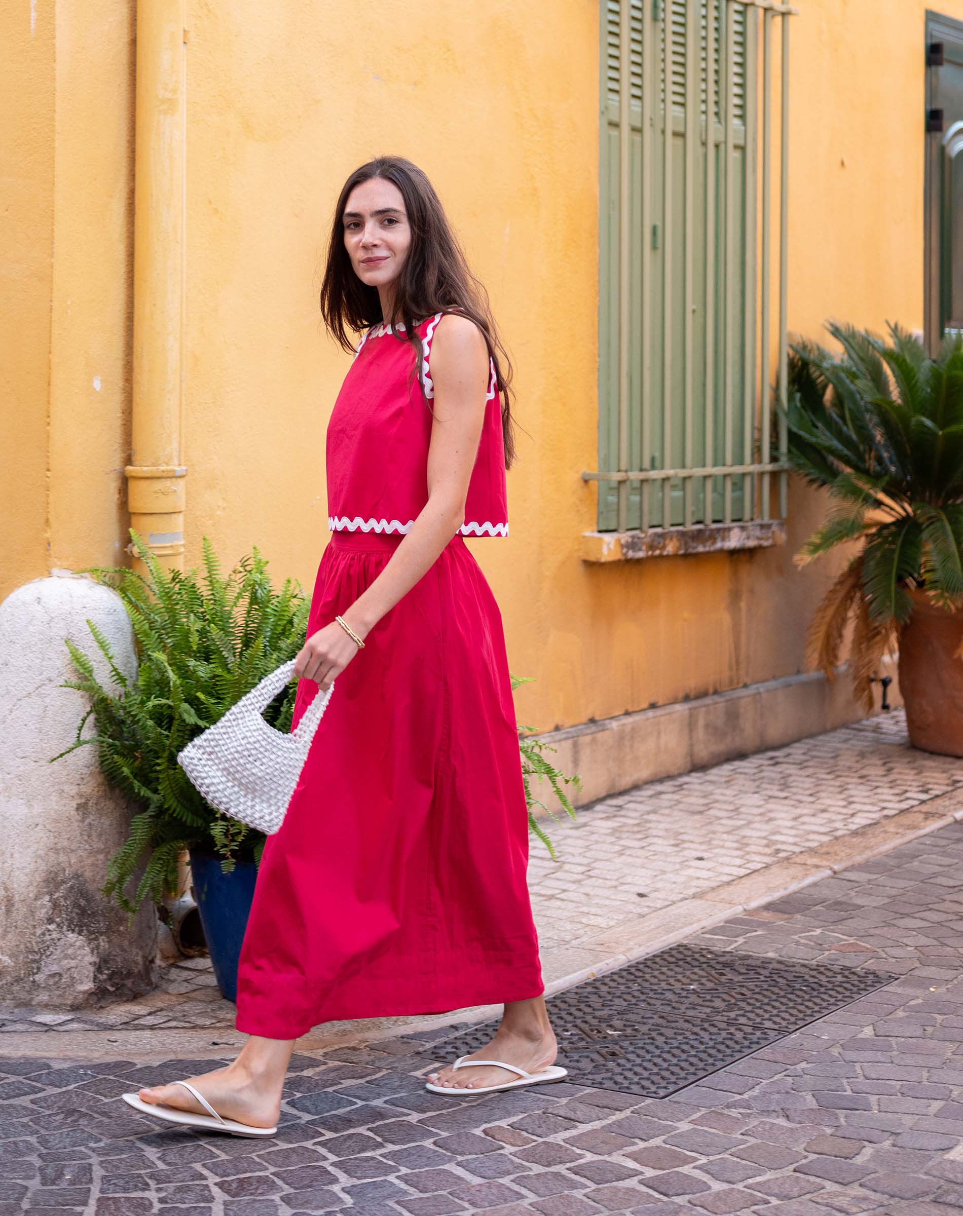 Woman in a red dress standing on a street with a yellow wall and plants in the background