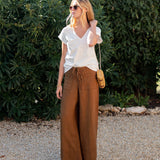 Woman wearing a white top and brown wide-leg pants standing outdoors with greenery in the background.