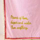 Pink blanket with chartreuse text  that says "oceans of love, deeper and wider than anything" with a natural tassel trim 