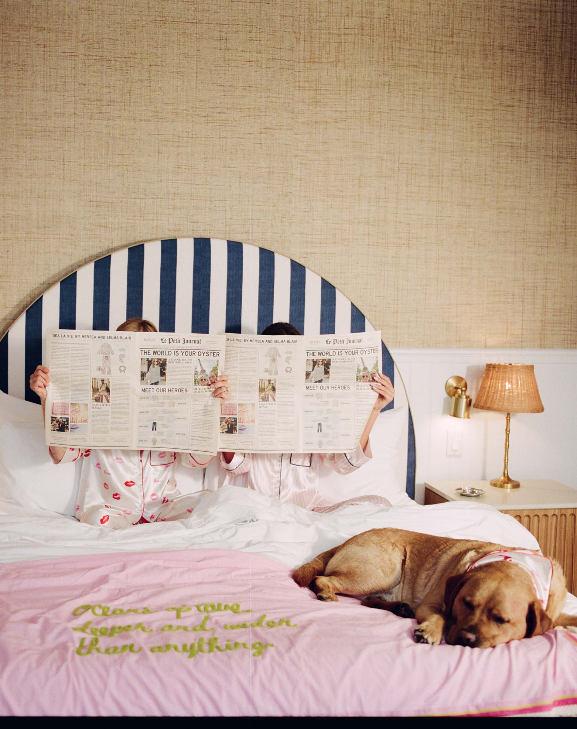Two people in bed with a dog, reading a newspaper, in a bedroom setting.