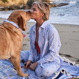 Woman sitting on a beach blanket with a dog, kissing it on the beach.