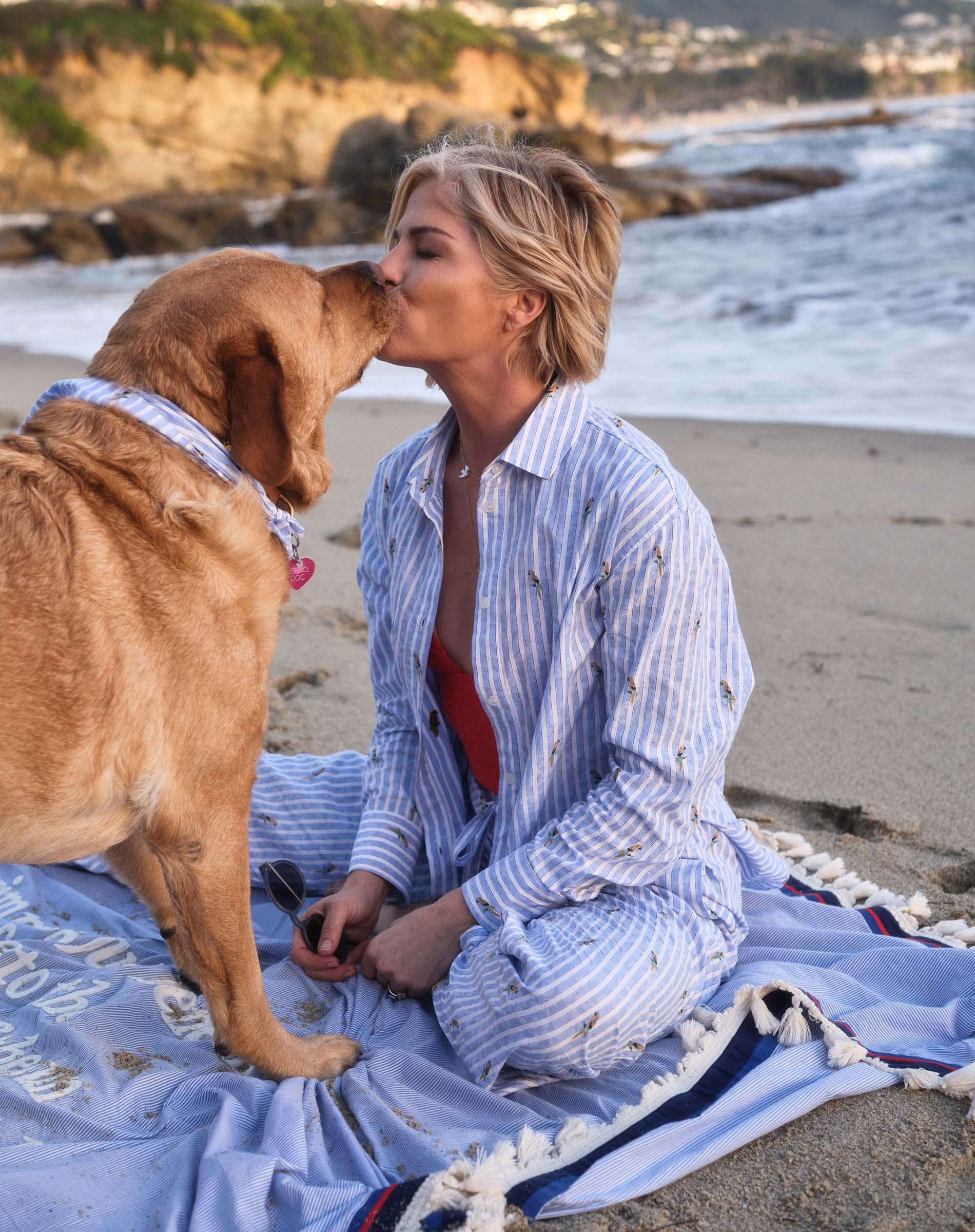 Woman sitting on a beach blanket with a dog, kissing it on the beach.