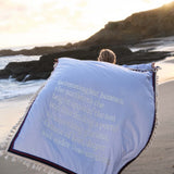 Person on a beach with a towel featuring a quote, with ocean and rocks in the background