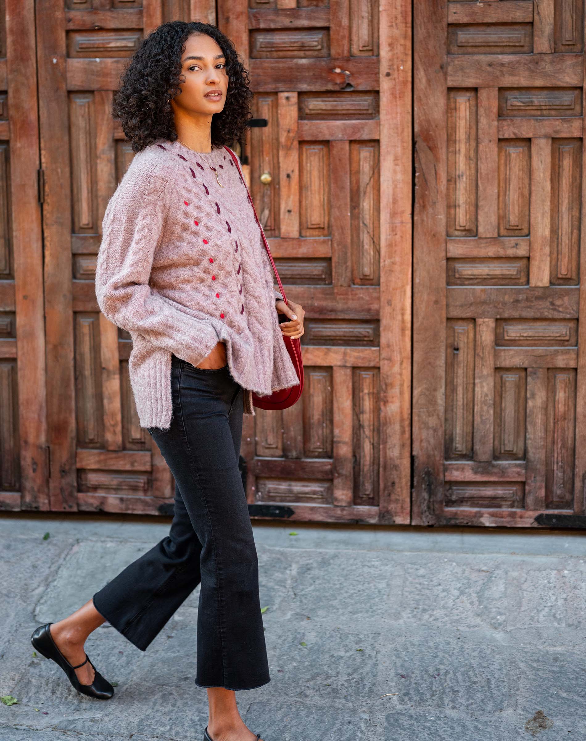 Woman in a pink knit sweater and black pants standing in front of a wooden door.