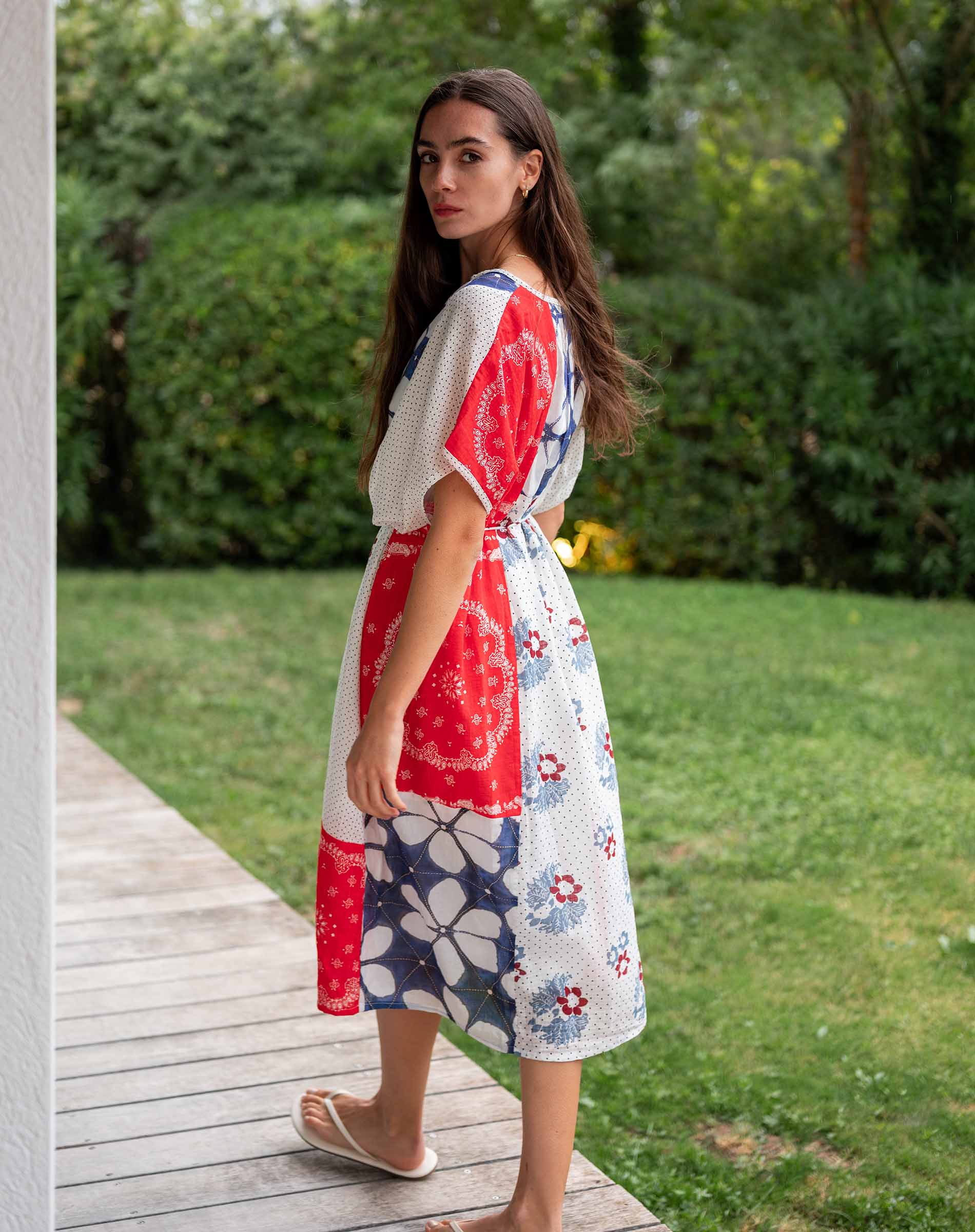 Woman wearing a red, white, and blue floral dress standing on a wooden path with greenery in the background