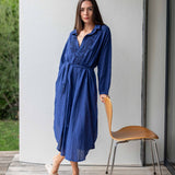 Woman in a blue dress standing next to a wooden chair on a wooden deck.