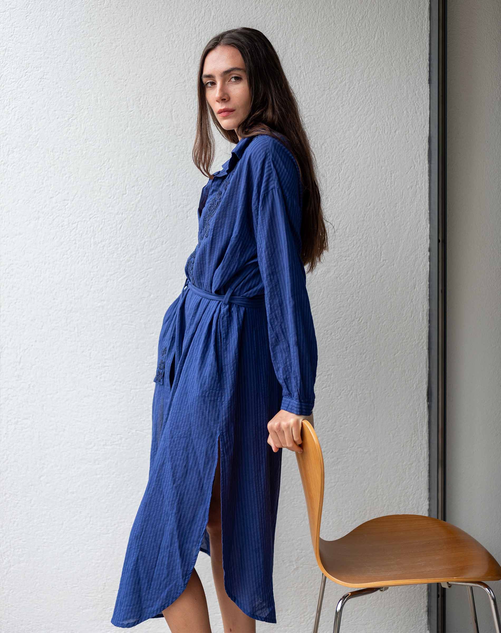 Woman wearing a blue dress holding a wooden chair against a white wall.