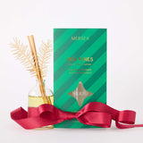 MERSEA women's travel-friendly sea Pines room diffuser in a clear glass bottle with wooden reeds and gold accents, accompanied by its green striped packaging and a decorative red ribbon. Text: MERSEA SEA PINES gardenia | cedar | santal ROOM DIFFUSER APPLIED TO SCENTS, MERSEA, 5 FL OZ.