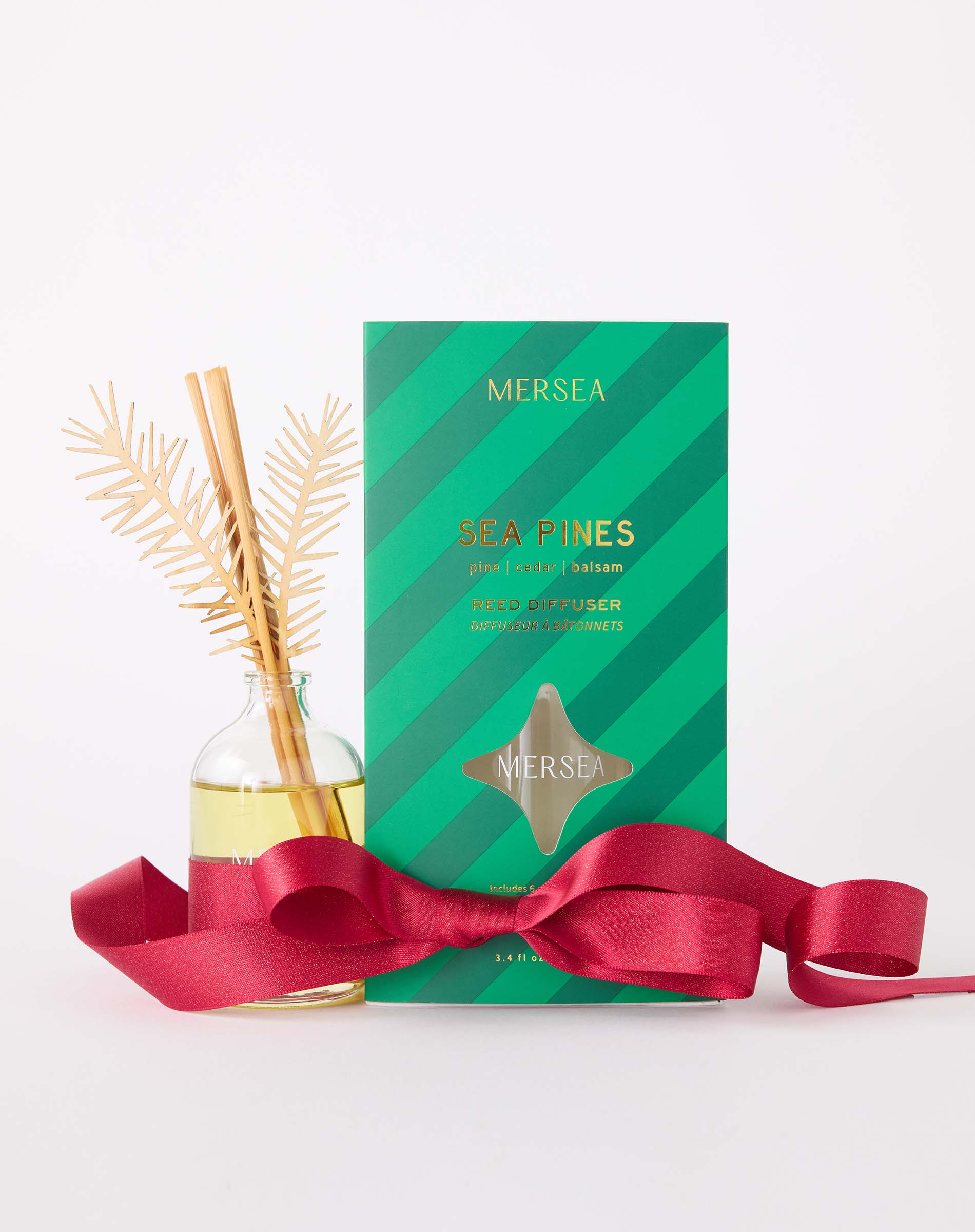 MERSEA women's travel-friendly sea Pines room diffuser in a clear glass bottle with wooden reeds and gold accents, accompanied by its green striped packaging and a decorative red ribbon. Text: MERSEA SEA PINES gardenia | cedar | santal ROOM DIFFUSER APPLIED TO SCENTS, MERSEA, 5 FL OZ.
