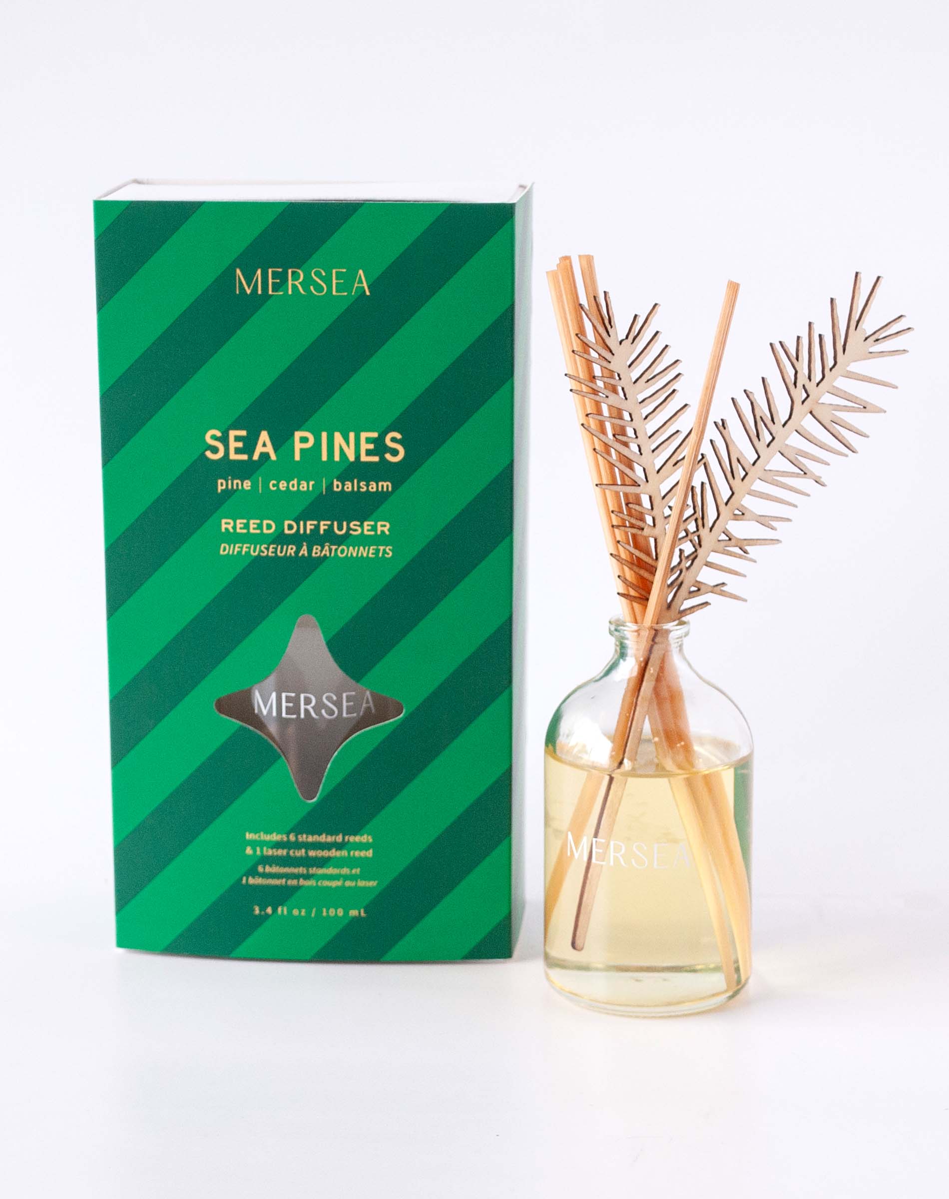 Sea Pines reed diffuser in a clear glass bottle with gold-toned sticks and a green striped box featuring the text: 'MERSEA SEA PINES pine | cedar | balsam REED DIFFUSER DIFFUSEUR à BATONNETS MERSEA'. Included additional details at bottom of box are "Inclus de: s tralxated results, 2 Pieces of scented wood, 4 dimensions: 15cm x 10cm x 10cm, Offer included in current promotion" and size '3.4 FL OZ / 100 mL'.