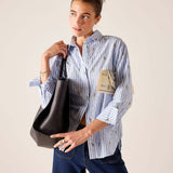Woman holding a black handbag and wearing a striped shirt with a beige bag over her shoulder against a plain background