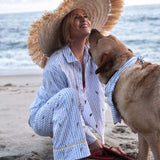 Woman in a straw hat and light blue outfit sitting on the beach with a dog.
