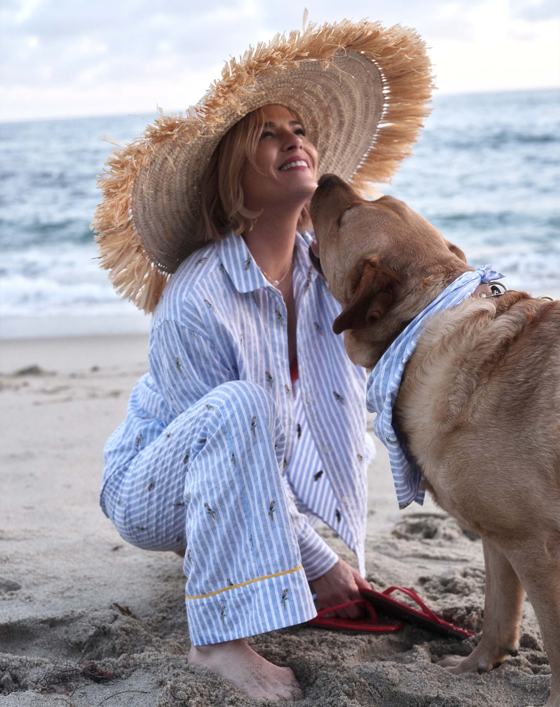 Woman in a straw hat and light blue outfit sitting on the beach with a dog.