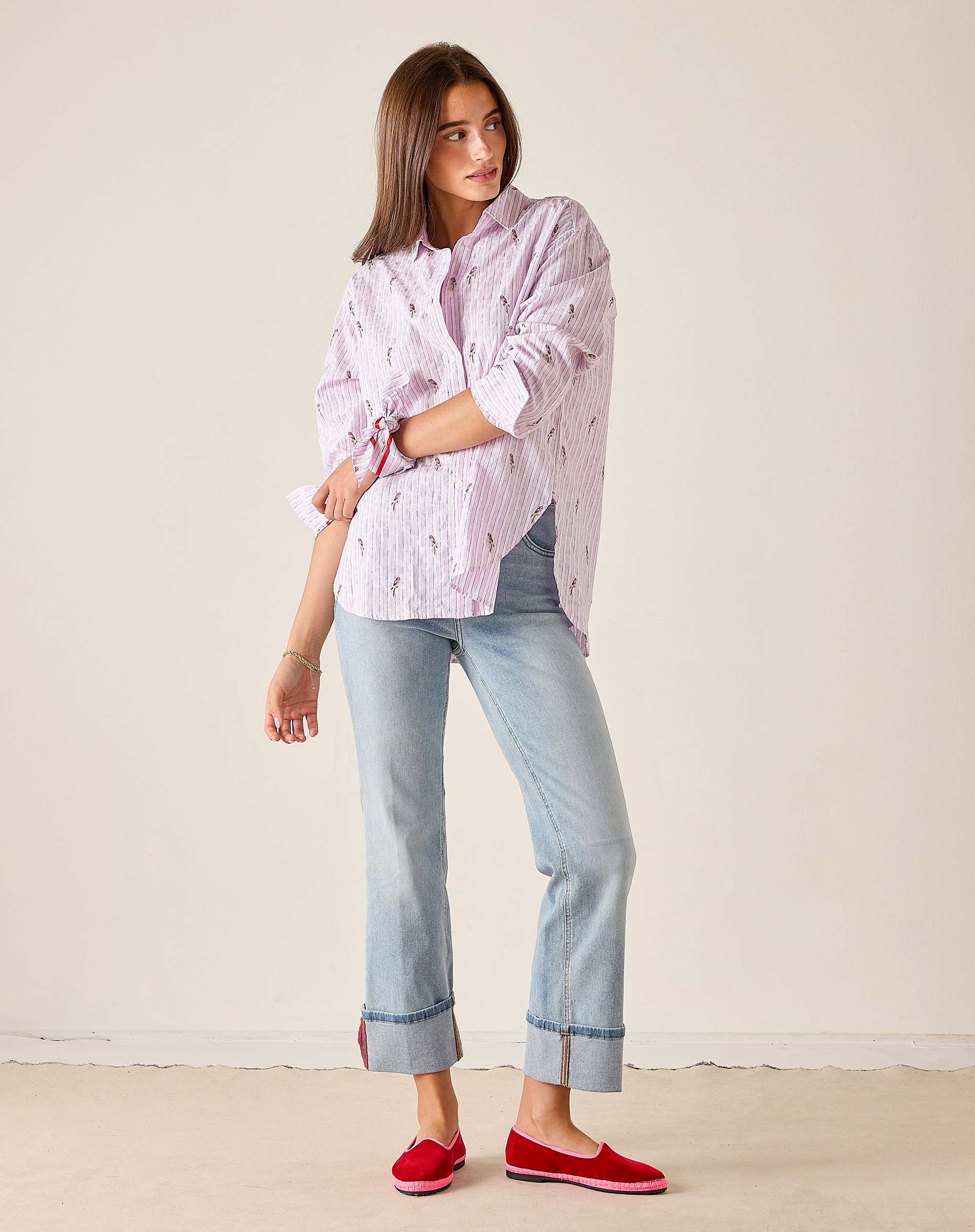 Woman wearing a pink striped shirt, light blue jeans, and red shoes against a beige background