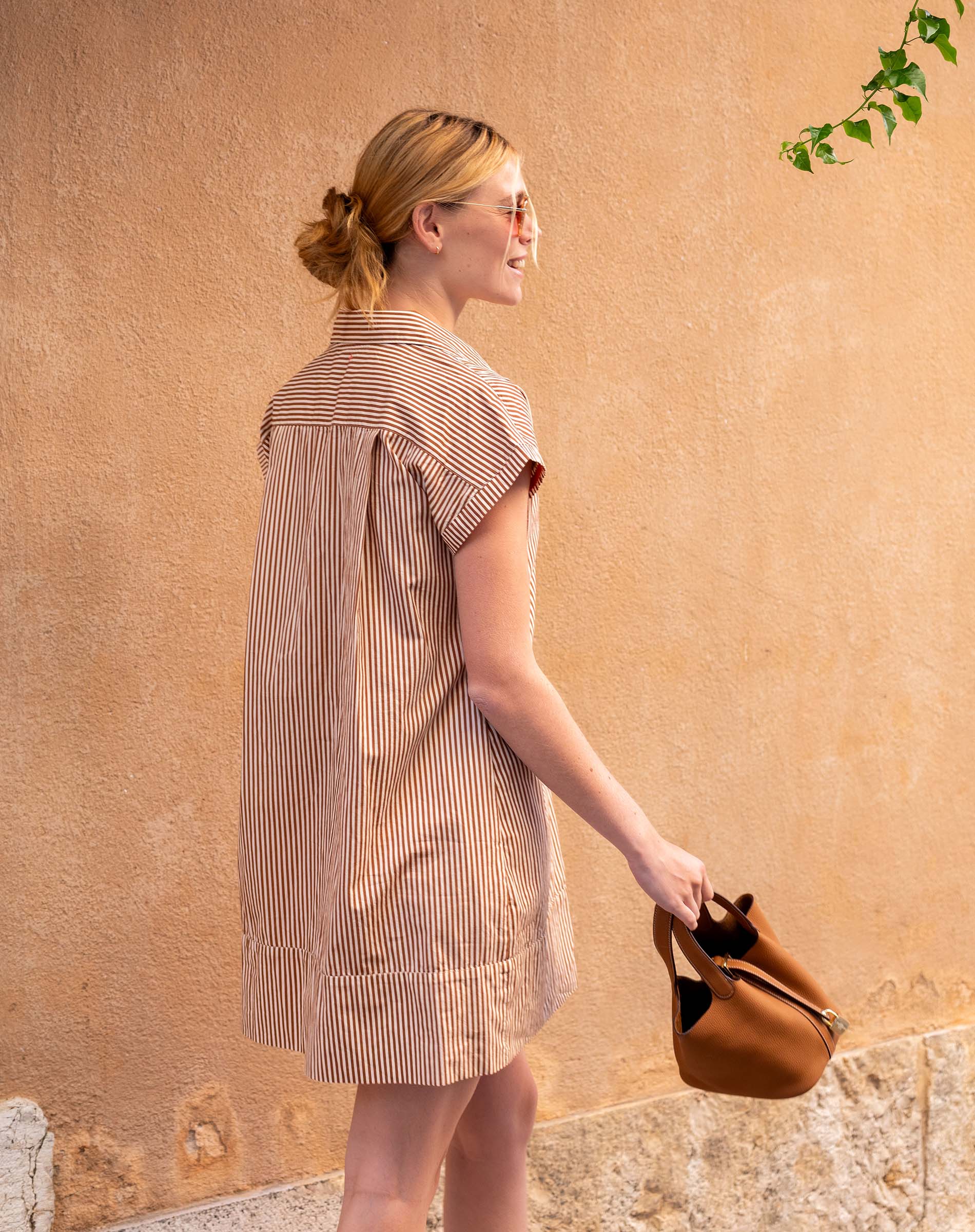 Woman in a striped dress holding a brown bag against a beige wall.