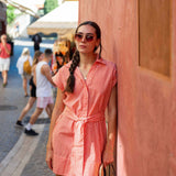 Woman in a red and white striped dress standing against a wooden wall with people walking in the background.
