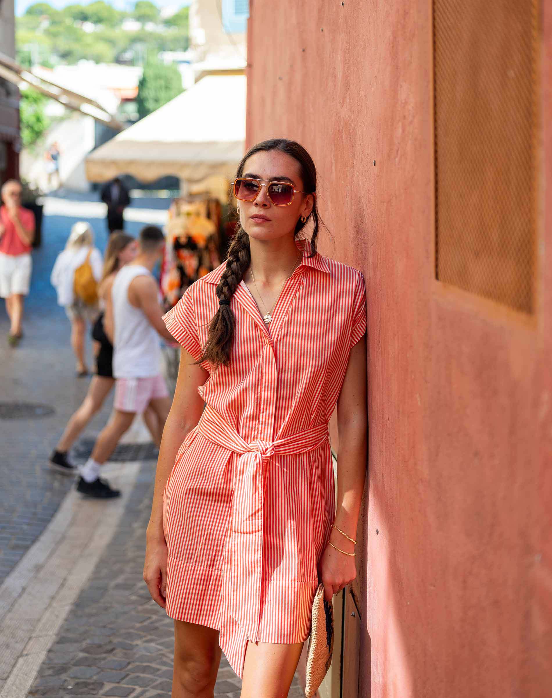 Woman in a red and white striped dress standing against a wooden wall with people walking in the background.