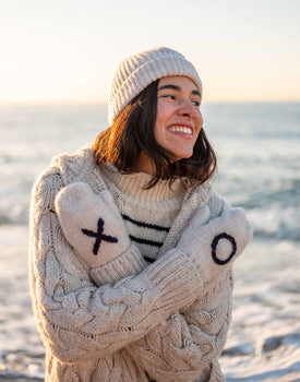 woman wearing mersea sailor love xo mittens in oatmeal and oxford blue with arms crossed in front with water in background