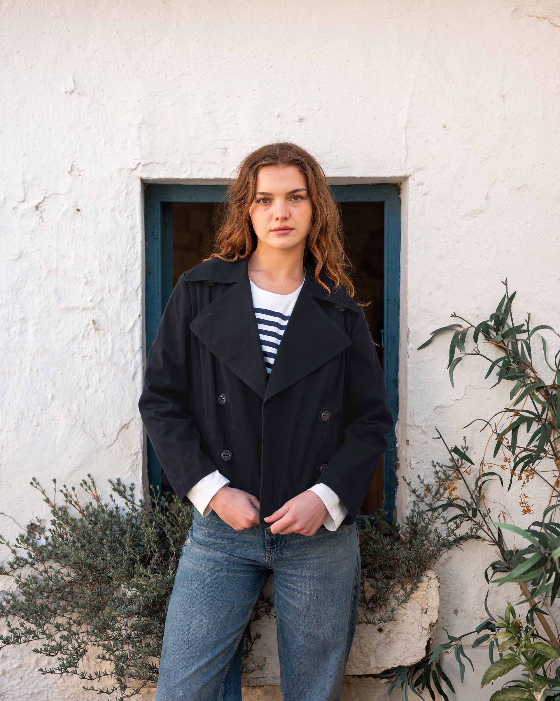 female wearing navy peacoat over white and blue striped tee shirt with blue jeans on white wall