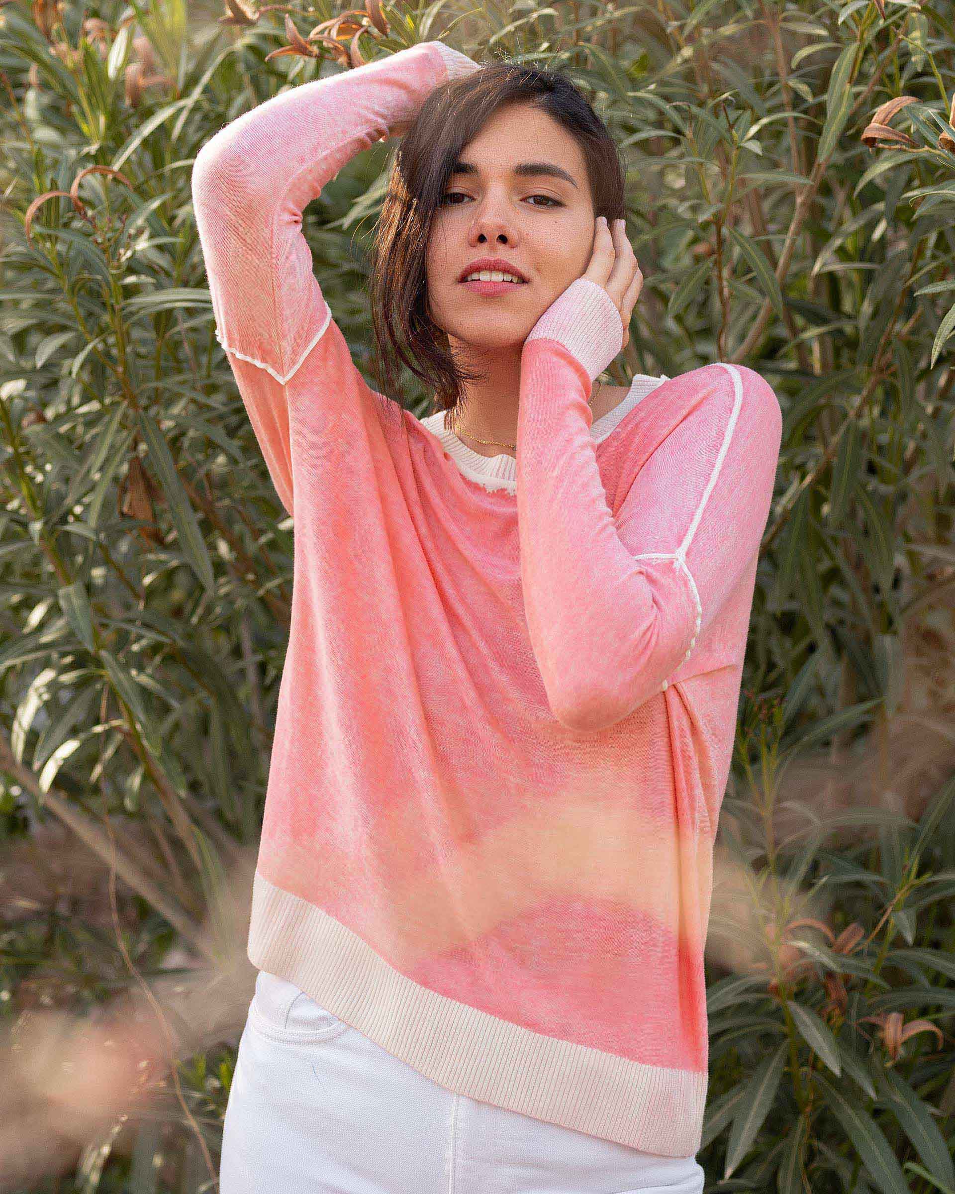 Female wearing pink hand-dyed cashmere sweater with hands behind her head stands in front of plants