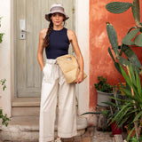 Woman standing in front of a door with plants around, wearing a navy top, white pants, and a beige hat.