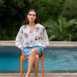 Woman sitting on an orange chair by a pool with a floral blouse and denim shorts.