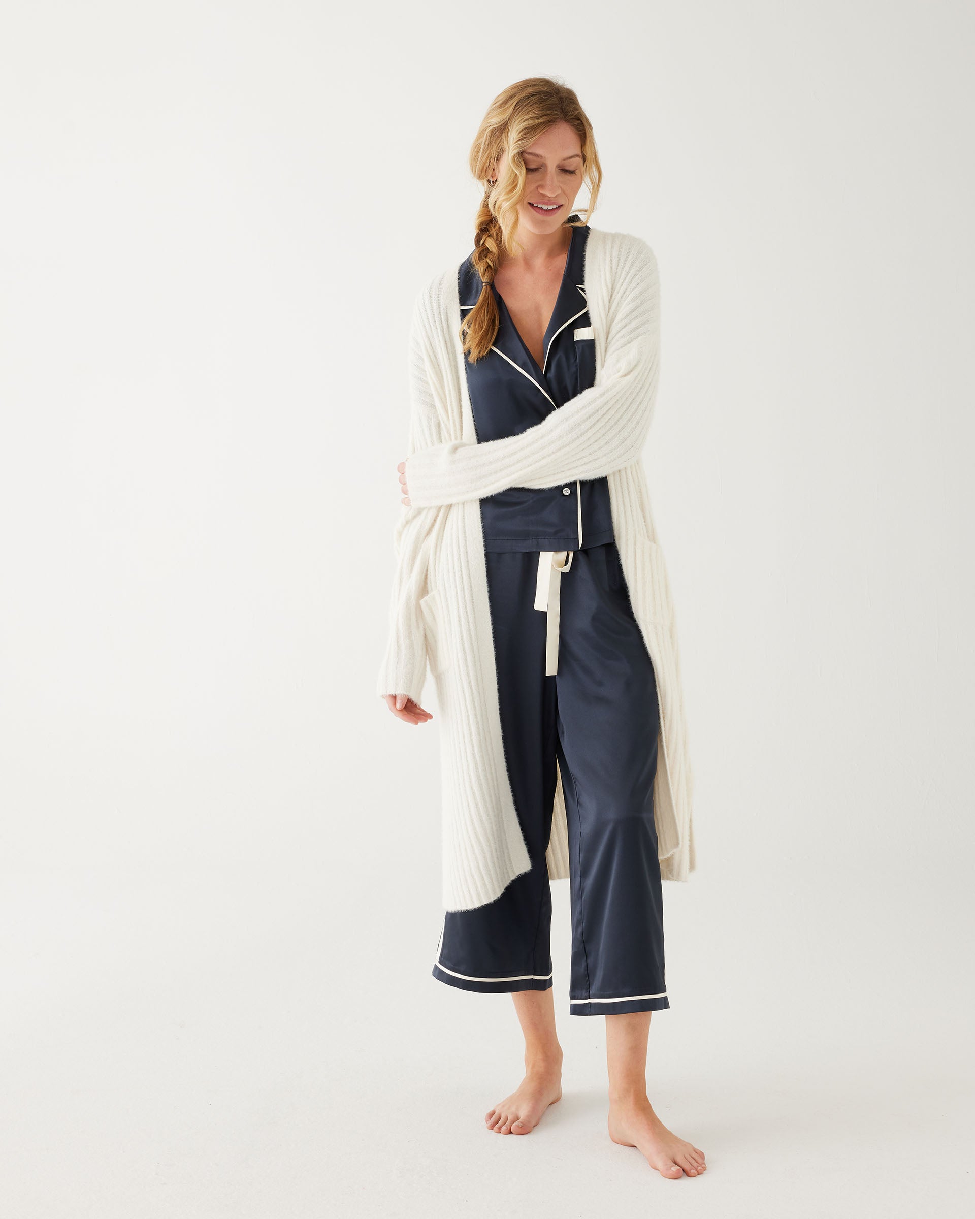 female wearing Mersea sapphire satin sailor pajama set and white robe standing against white background