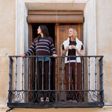 Two women standing on a balcony with an ornate iron railing, looking at each other and smiling. One woman wears a striped knit sweater in truffle and sky colors, while the other wears a cream wrap over a dark top.