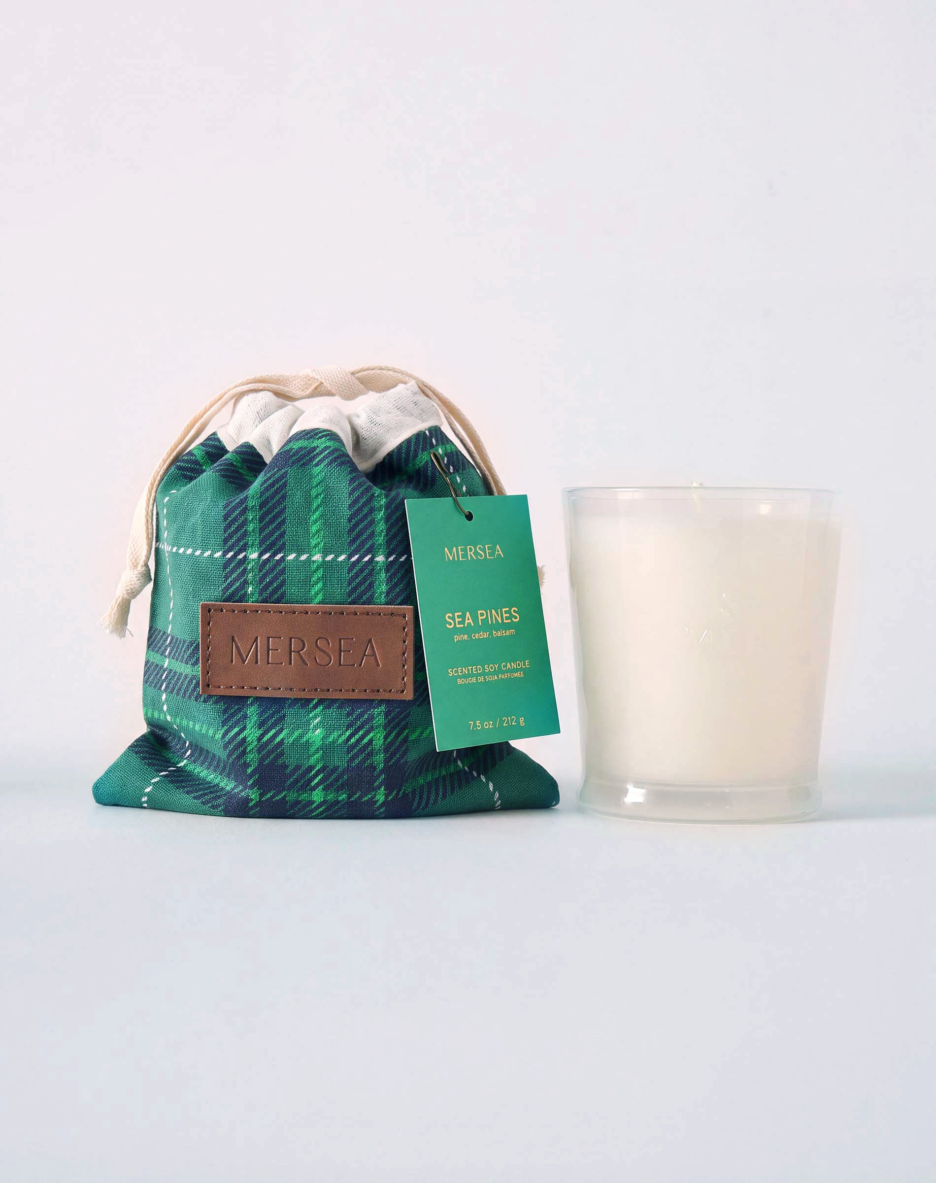 MERSEA women's travel-friendly green and navy plaid drawstring pouch bag and a white candle in a clear glass jar with a green label that reads 'MERSEA SEA PINES'. Text: "MERSEA SEA PINES, SEA PINE SCENT, MERSEYED SOY CANDLE, HAND POURED IN THE USA 10 OZ / .32 OZ""