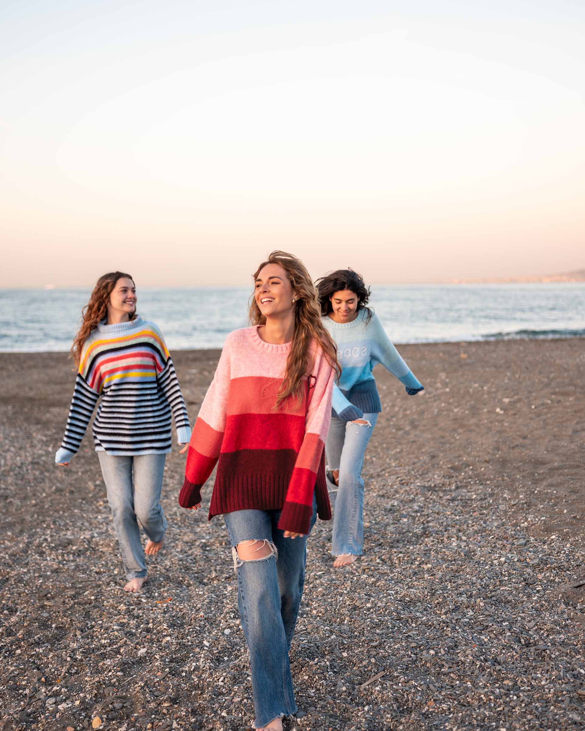 women wearing mersea seahappy heart peace and rainbow holiday sweaters in bliss pink stripes and galxy blue stripes on beach