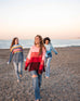 women wearing mersea seahappy heart peace and rainbow holiday sweaters in bliss pink stripes and galxy blue stripes on beach