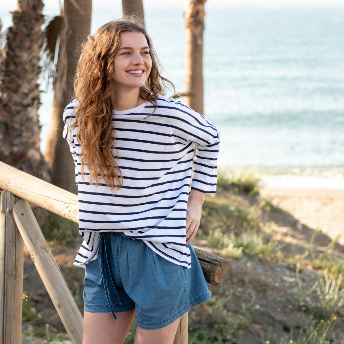 female wearing a navy striped tee shirt leaning on a fence by the beach 