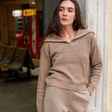 Woman wearing a beige sweater and pants standing against a wall.