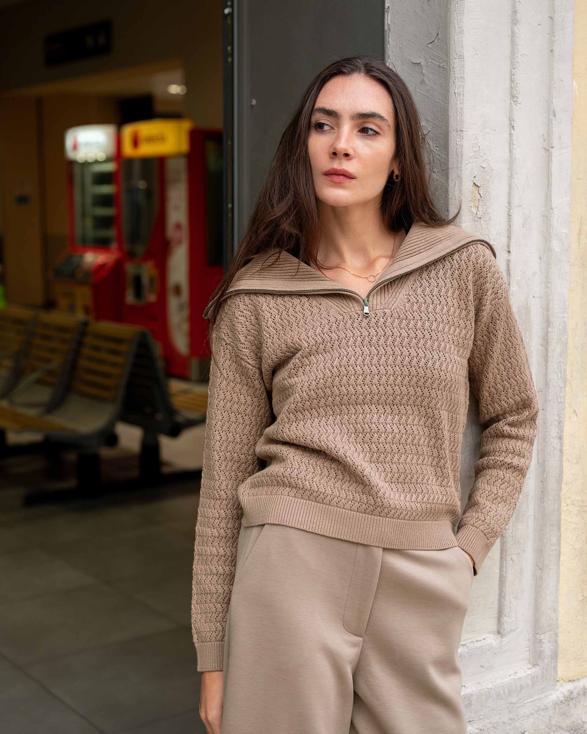 Woman wearing a beige sweater and pants standing against a wall.