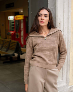 Woman wearing a beige sweater and pants standing against a wall.