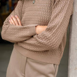 Person wearing a beige knit sweater and pants leaning against a wall.