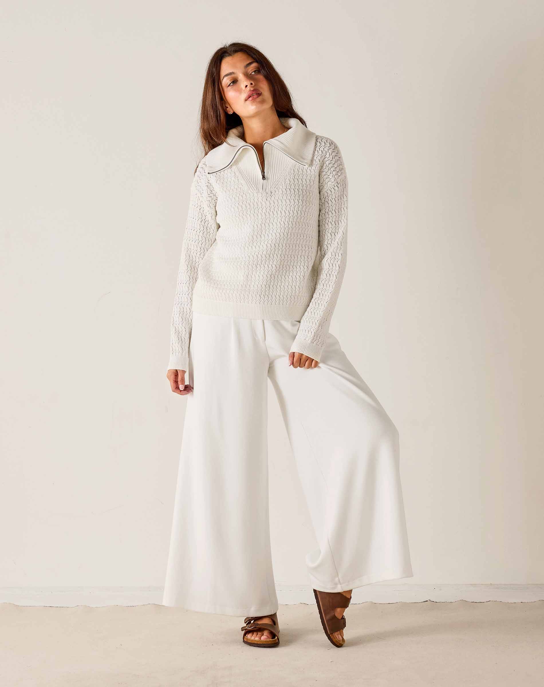 Woman wearing a white knit sweater and wide-leg pants against a plain background