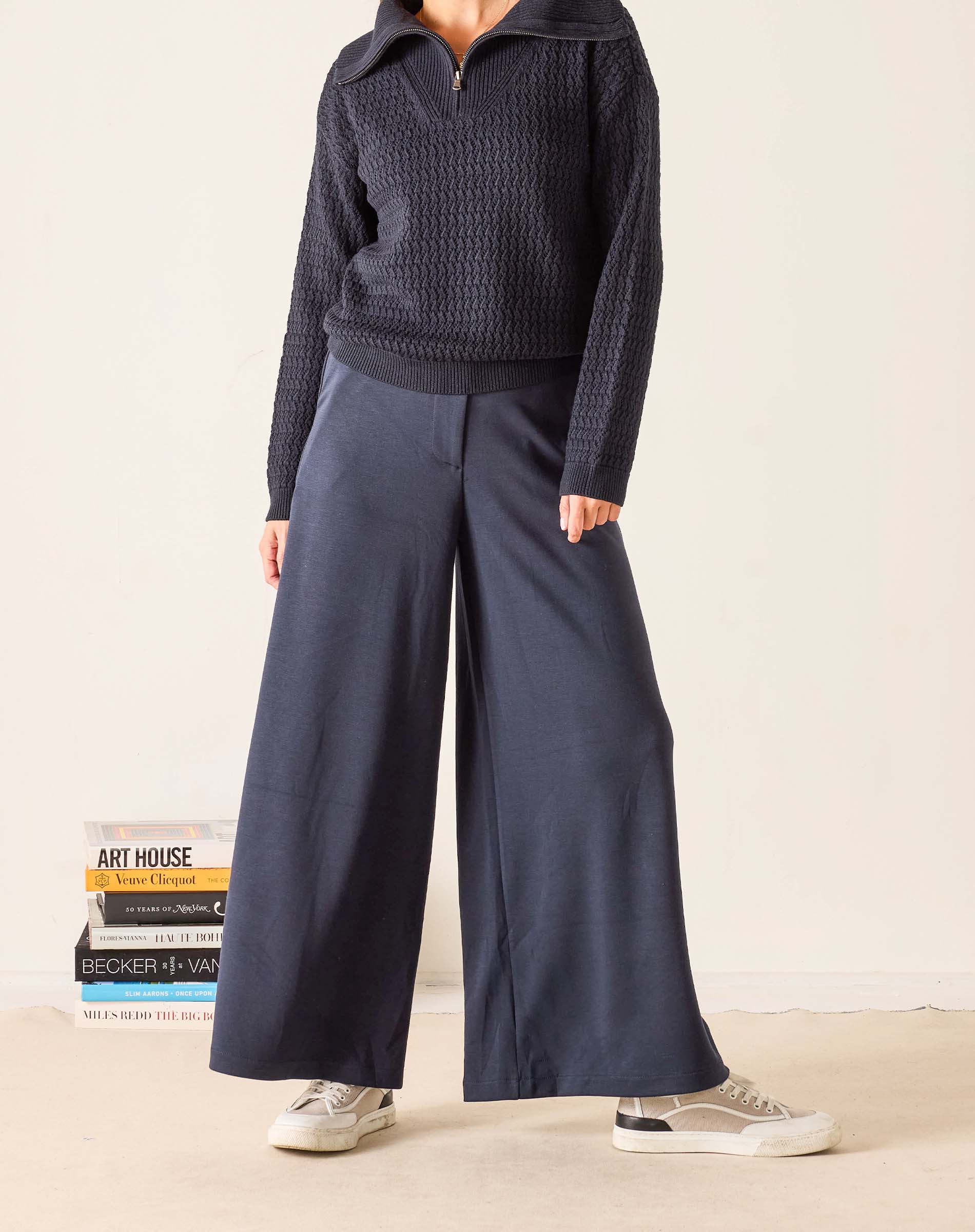 Person wearing a dark sweater and wide-leg pants standing next to books on a light background