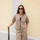 Woman in beige outfit with sunglasses holding a suitcase against a light-colored wall.