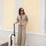 Woman in beige outfit with a suitcase against a light-colored wall.