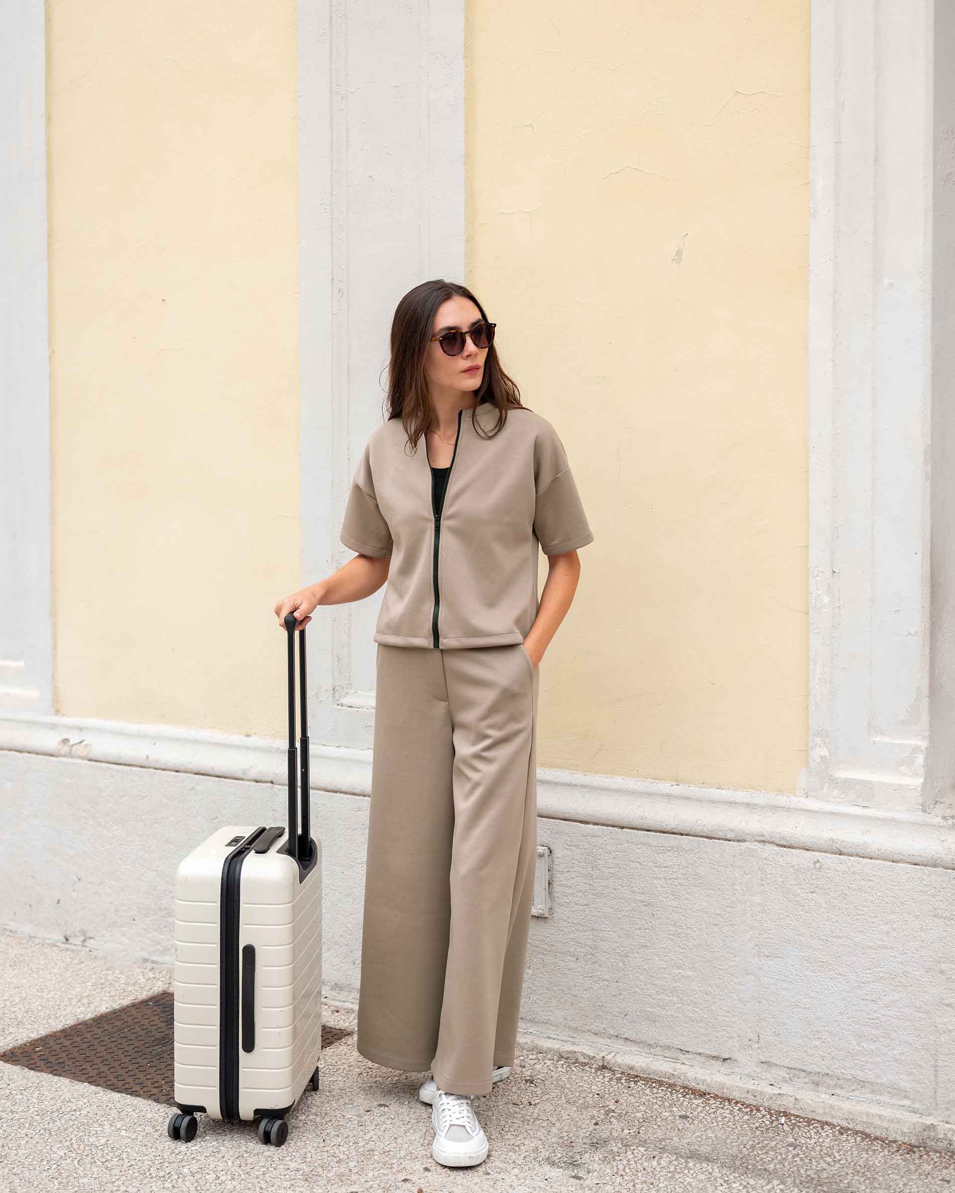 Woman in beige outfit with a suitcase against a light-colored wall.